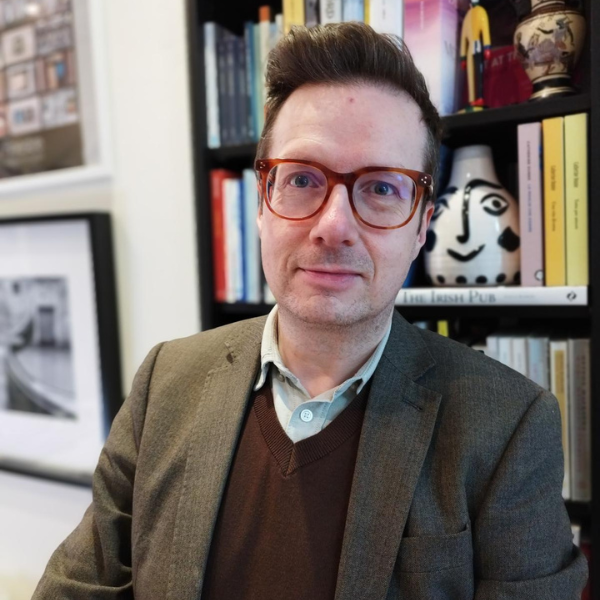 Middle-aged, white man with short hair and statement glasses, wearing a brown blazer, with a bookcase and some framed photographs in the background